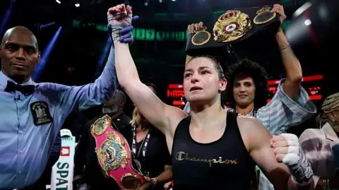 Katie Taylor, centre, with dark tied back hair, wearing a black vest with the word Champion on it. There is a man to her left wearing a blue shirt and a black bow tie, raising her right arm in the air.