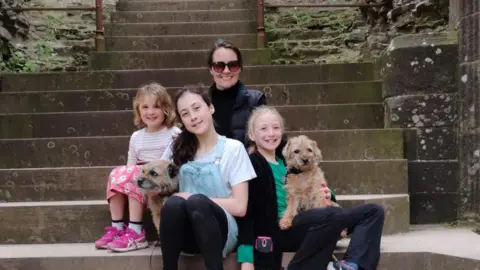 Handout Jenny Purshouse poses with her three children and dog on some stone steps. She is wearing a coat and dark sunglasses, and has dark hair. 
