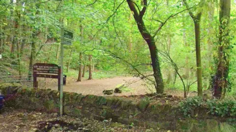 Google A park entrance with a wooden sign that reads Gore Glen Woodland Park with a pathway surrounded by green trees