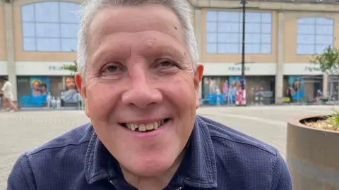 Nigel Brown is sitting on a bench in a square in Truro. He is wearing a blue T-shirt with a half-zip. He has short grey hair and is smiling broadly. To his left is a large concrete planter which has grasses in it. There are a few people walking across the square in the background and there is a Primark, a JD Sports and a Wetherspoons.
