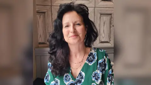 Anne Thackeray A woman with long, wavy dark hair. She wears a green top with blue flowers, and a gold necklace and earrings.