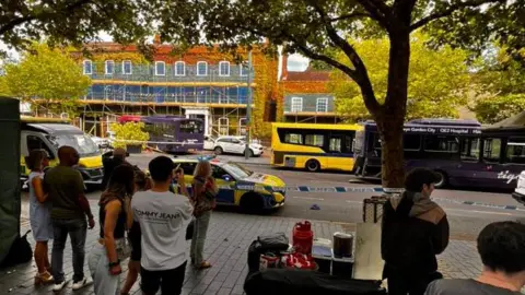 Elvio Abreu Pedestrians watch on from the pavement following the bus crash. The crashed bus can be seen in the distance behind a cordon with police cars parked up as well as other buses.