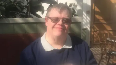 Neil Blackman wearing glasses sits at a marble café table with a cup of coffee, in a bright indoor space with plants and sunlight casting shadows on the wall behind.