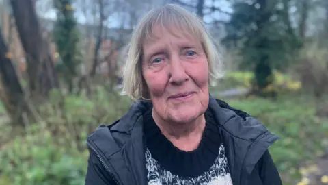 Elizabeth Baines / BBC A woman with white hair stands in a woodland. She wears a black jumper and black puffer coat