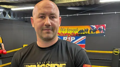 Jordan Doherty standing beside the boxing ring. He is bald and is wearing a black t-shirt with Bristol Ringside's logo on it.  In he background there are two posters on a black wall.