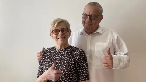 A smiling Val and Geoff King. She is wearing glasses and a black and white top. He is wearing a white shirt and both are holding their thumbs up in front of them. 