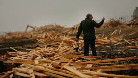Getty Images A man standing near a pile of timber on a pebbled beach.