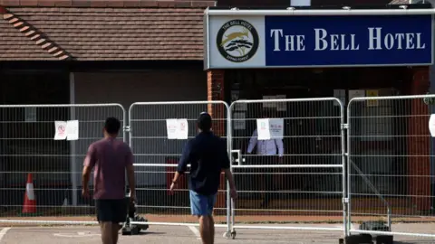 Reuters Two men in T-shirts and shorts walking with their backs to the camera towards The Bell Hotel.  There is a metal fence in front of the hotel building.