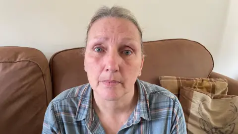 a middle aged woman is sat on a brown sofa with her hair tied back wearing a blue checked shirt.