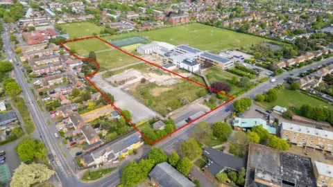 An aerial view of a school which has large playing fields either side. On the left, a playing field and car park is outlined with a red line. There are hundreds of homes around the school site.