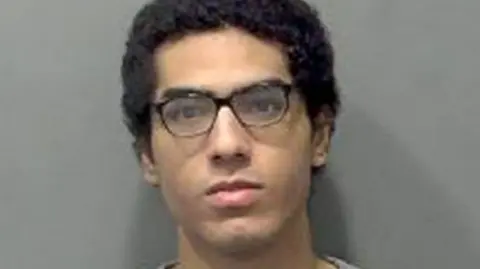 Nicholas Prosper, who has dark hair and dark glasses, staring into the camera for a police custody image. He is wearing a grey sweatshirt.