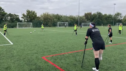 Six female amputee footballers standing in a circle on a pitch. Four are wearing fluorescent bibs over black t-shirts and shorts. All the women have crutches. Four training goals can be seen on the far side of the pitch.