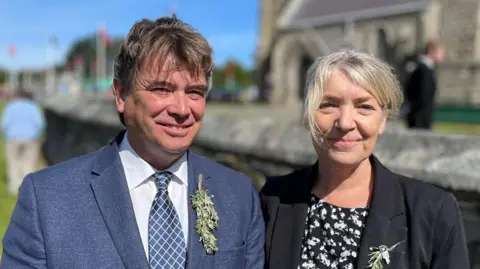 BBC Kirrie and Philip Jenkins smile looking at the camera outside the Royal Chapel in St John's. He is wearing a blue blazer with a white shirt and blue tie with a crisscross pattern in white and a sprig of bollan bane in his button hole. She is wearing a black jacket over a black top with a white floral pattern on it and also has bollan bane pinned to her lapel.