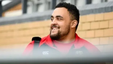 Anthony Nolan Ellis Joseph is wearing a red and black team top as he looks over his shoulder, smiling behind him