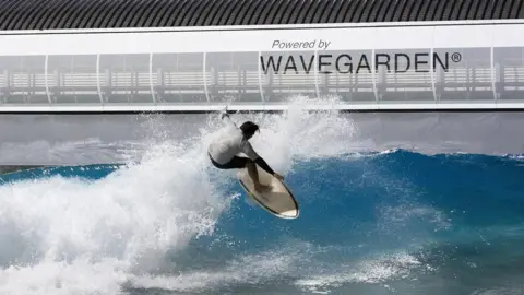 The Wave A man on a surfboard rides a wave at The Wave in Bristol. The water is a blue-green colour with white foam, and he is wearing a white top and black shorts