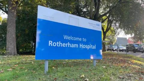 A large blue hospital sign on the grass outside a hospital with a large car park