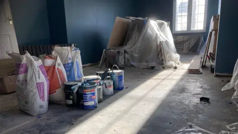 A room inside Anna Mimms's home has paint and bags of cement on the floor. The work to repair the home after Storm Babet has not yet been completed. The picture shows a shaft of light, shining through the a window