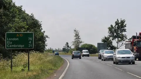 J.Hannan-Briggs/Geograph The A10 between Littleport and Ely. It is a single carriageway road with traffic on both sides. On the left in the verge is a green distances sign saying A10 with London, 76, Ely, 4, and Cambridge, 21, listed on it. 