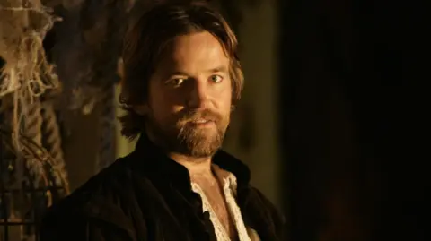 Head and shoulders shot of Dean Lennox Kelly playing Shakespeare in BBC drama Dr Who. He has a beard, a collar-less black jacket and white shirt and is in a dark room