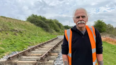 Neil stands in front of the new section of track being laid for the project. He has long light grey hair and a large grey moustache, and is wearing a black fleece with a bright orange high-vis vest over the top. 