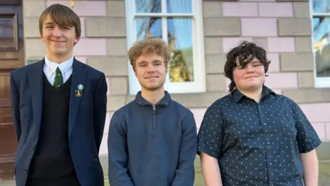 Alaric, Oliver and Ashley are standing side by side in front of a building with pink and stone brickwork. Alaric is wearing a dark suit with a tie and badge, while the others are dressed in casual tops. A window and part of a wooden door are visible behind them.