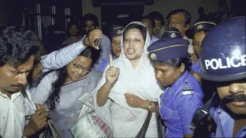 Getty Images Khaleda Zia is wearing a white headscarf and saree and gesturing with a raised finger, while she is taken away in a crowd by a group of police officers in blue uniforms and hats