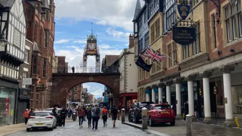 Generic view of Chester city centre showing the city wall and clocktower with pedestrians walking along the cobbled street.