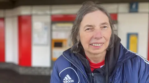 Paula Mitchinson is smiling against the red and white backdrop of the Stadium of Light metro station. She is wearing a blue Adidas puffer jacket with Sunderland AFC's crest and has short grey hair.