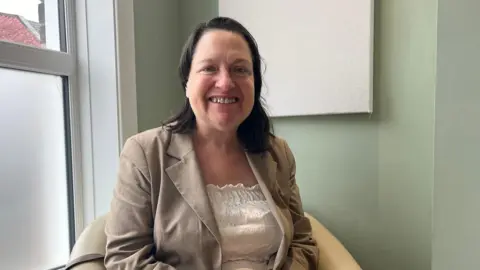 Clare Worden/BBC Alexandra Kemp smiles at the camera as she sits in a cream-coloured chair. She is wearing a cream jacket and top. Her hair is worn in a dark brown bob. 