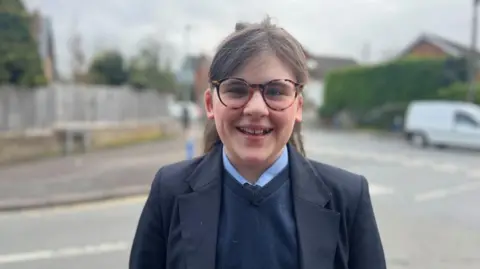 A young girl with brown hair and multi-coloured round glasses, wears a navy blazer and jumper, with a pale blue shirt. She is smiling at the camera while standing in front of a blurred road junction. There is housing, hedges and a white van in the distance. 