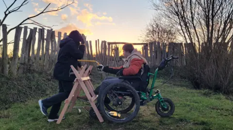 Two young people outside. One is stood up in a black coat and a hood, their face is covered. The other is in a wheelchair and wearing an orange and beige jacket. They are in a woodland setting with trees and logs positioned around them. The sun is setting. 