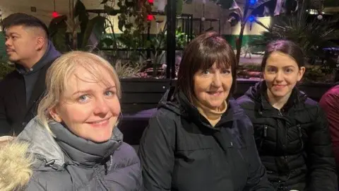 Three women smile. They are sitting down and wearing coats. 