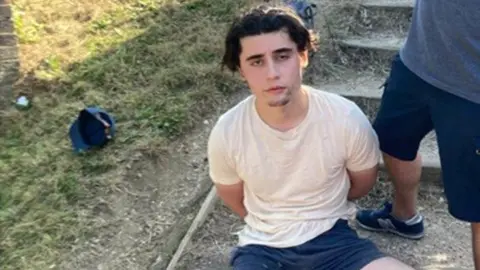 Handout photo issued by the Metropolitan Police of Daniel Khalife taken after his arrest on a canal towpath on 9 September 2023 - he has medium length hair and is looking at the camera. He is wearing a cream t-shirt and blue shorts, and looks to have his hand handcuffed behind his back. 