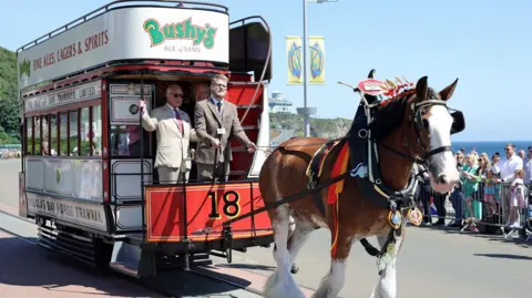 The King, wearing a beige suite and sunglasses, standing at the front of a horse drawn tram.