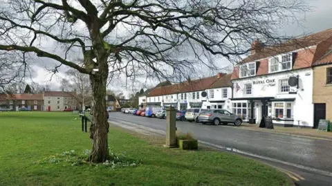 Google A view of Great Ayton high street with the Royal Oak hotel and a line of white shops with cars parked in front of them on the right and a village green on the left.
