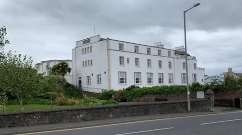 A white fronted hotel with a large number of windows behind a low stone wall and trees with the sign North West Castle Hotel almost obscured by plant life