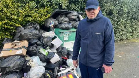 Teddy Smith berdiri di sebelah Black Bin Bags di jalan di sebelah tempat sampah industri yang meluap. Dia mengenakan bulu biru, bagian bawah pakaian olahraga biru dan topi biru.