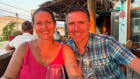 Emma Carnell A woman and a man sat side-by-side looking towards the camera. They are sat at a table with wine glasses in front of them. 