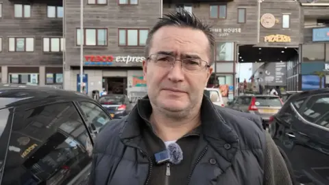 A man stands in a car park in front of his electric car, which is charging up. 