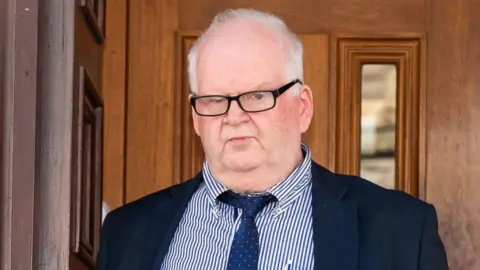 David Baxter is wearing a blue and white stripped shirt with a navy blazer and tie. He has short grey hair and black glasses on. Behind him is a wooden door. 