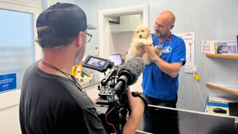 Paul Crompton Film-maker Danny MacGregor working behind the camera in a veterinary clinic. His back is to the camera. TV vet and animal welfare campaigner Dr Marc Abraham OBE is in front of him holding a dog. He is smiling. There is a stethoscope around his neck.