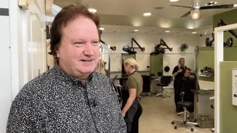 Adrian Ball smiles and looks off to the right of the image. He's wearing a black and white shirt, in a 'dotty' formation. Behind him people are sat in hairdressing chairs getting their hair done.