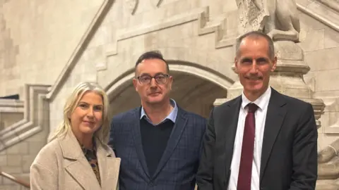 Christine Lock with blond hair wearing a cream coat (left) with husband Richard (centre) with brown hair wearing a pale blue shirt navy jumper and blue suit jacket and MP Bill Esterson with grey hair wearing a navy suit and burgundy tie. They are all smiling.