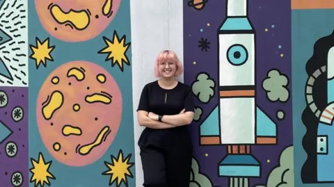 Dr Eleanor S Armstrong A woman wearing all black, with her arms crossed and smiling, in front of a white, blue and purple animated background full of shapes.