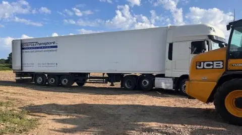 Sue Tyler-Moore A very long white lorry pulled up alongside a yellow telehandler in a field. The rear doors of the lorry are open.