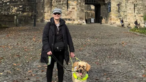 Terri Ballon smiles as she stands with her yellow Labrador guide dog on a cobbled path leading up to the stone gateway of a medieval castle behind her. She wears a grey cap and black shades, along with a black coat, top and trousers.