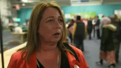 Kerrie is wearing an orange shirt with a Labour rosette and has long, brown hair. She is being interviewed at an election count.