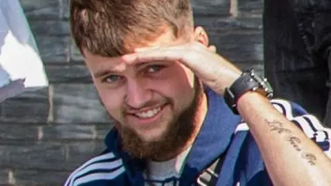 Conner Marshall, wearing a blue tracksuit jacket and a grey t-shirt, smiles as he holds his hand over his eyes in the sunshine