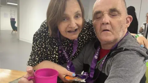 Sense A woman with short brown hair is standing next to a man wearing a grey hoodie. They are smiling while holding medals around their necks.