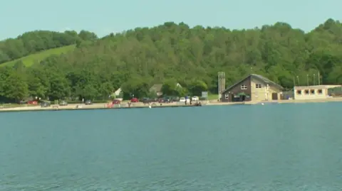 A wide outdoor scene with a large body of calm water in the foreground.
Across the water, there is a shoreline with several buildings and a small parking area. On the left side, there is a large modern building with a sloped roof and wide windows. To the right, there is a smaller building with a pitched roof and light-colored walls, and another low building beside it. A number of parked cars are lined up along the edge of the water, and a few small figures can be seen walking or standing near the shoreline. Behind the buildings, the background is filled with dense green trees covering rolling hills.